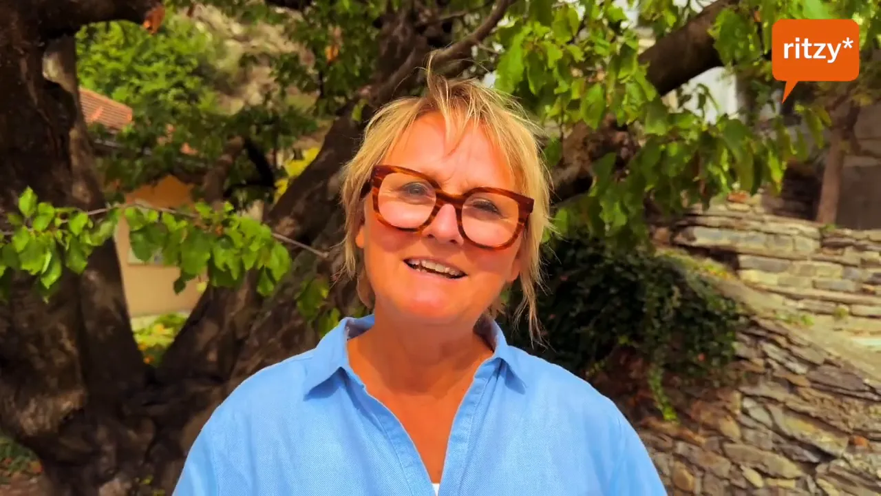 Frontalaufnahme einer lächelnden Person in hellblauer Bluse vor einem Baum und einer Steinmauer, ritzy*-Logo oben rechts