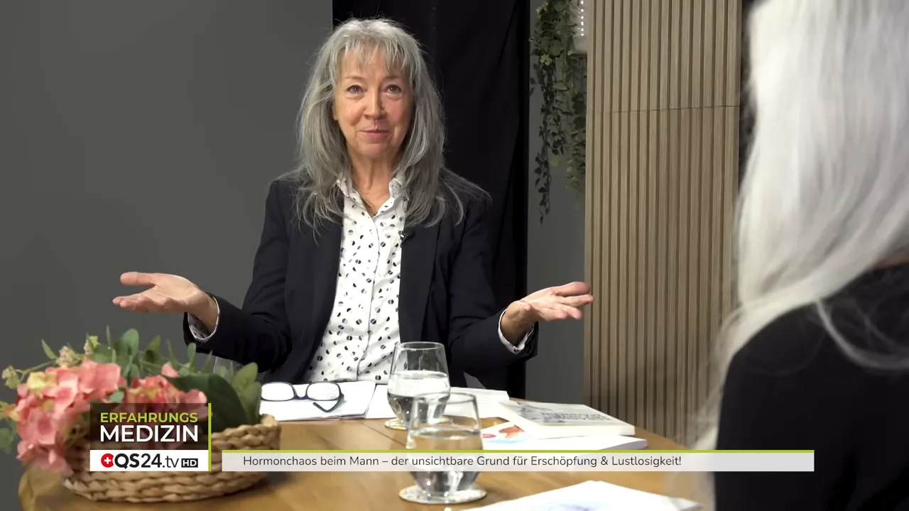 Expertin gestikuliert mit offenen Händen im Interview vor Tisch mit Glas und Unterlagen