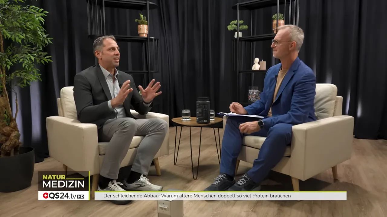 Weite Studioaufnahme mit zwei Gesprächspartnern, Produkt steht deutlich sichtbar auf dem Beistelltisch zwischen ihnen