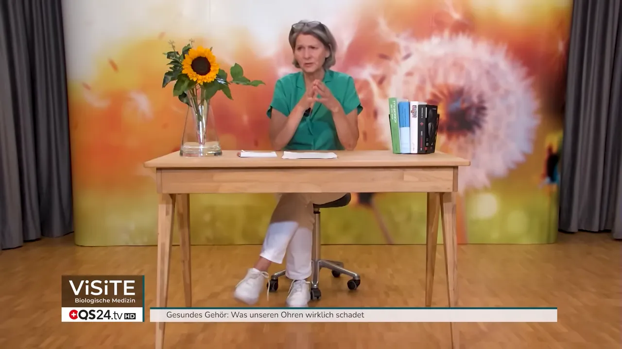 Weitwinkelaufnahme von Dr. Petra Wiechel am Tisch im Studio mit Sonnenblume und Büchern, gestikulierend