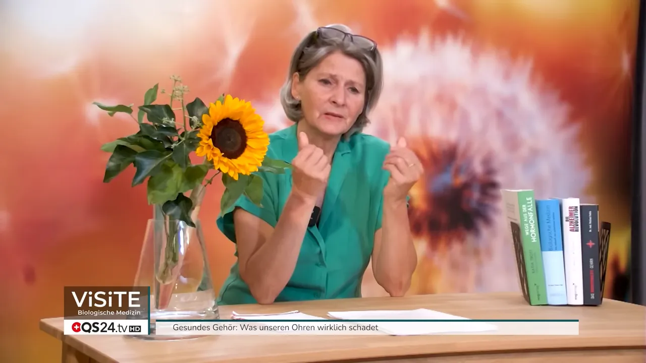 Dr. Petra Wiechel erklärt gestikulierend am Tisch (Handbewegung wie eine kleine Pinzette), mit Sonnenblume und Büchern im Bild — passend zur Empfehlung, Ohren nicht zu stark zu reinigen.