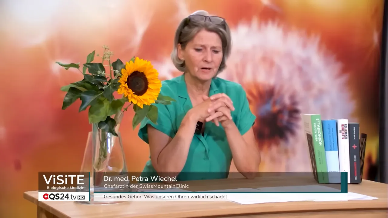 Dr. med. Petra Wiechel am Tisch im Studio mit eingeblendetem Namensbalken, Sonnenblume und Bücher im Vordergrund