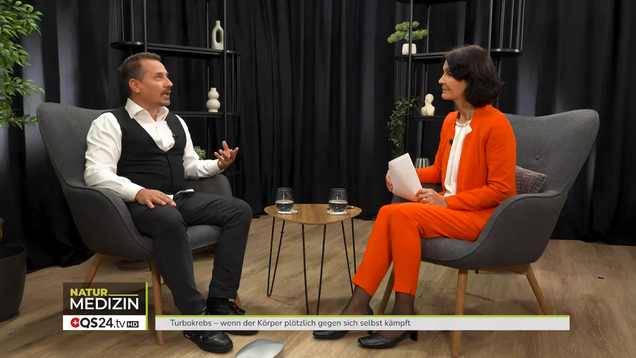 Zwei Personen im Studio-Interview am Tisch, Moderatorin und Gast im Gespräch