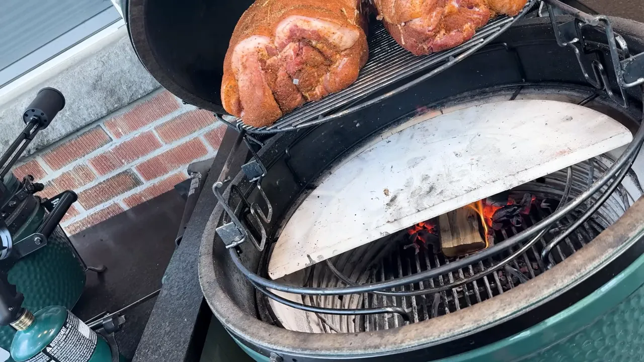 Open Big Green Egg with two pork shoulders on an elevated rack above a rear deflector and small wood fire