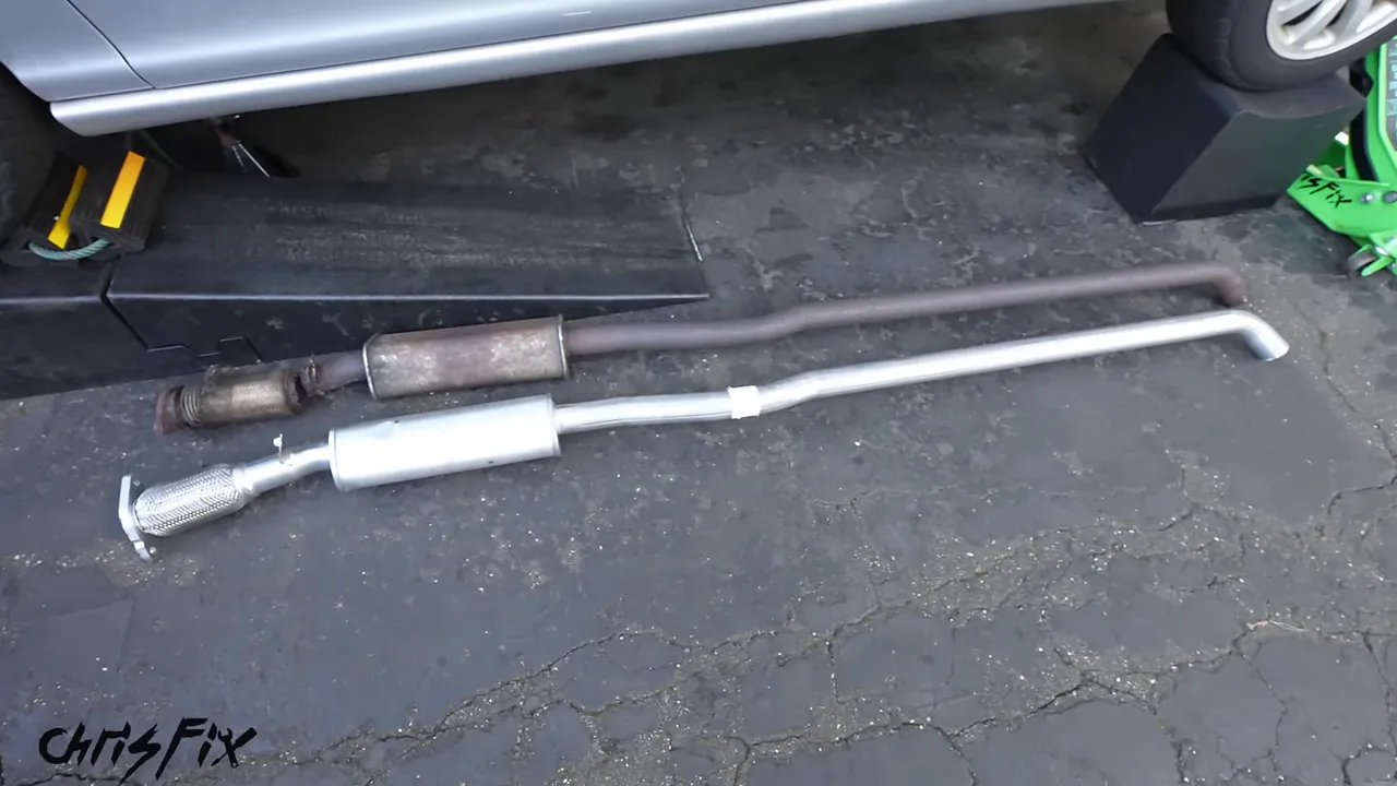 Old rusty exhaust section and new silver replacement section lying side by side beside the car