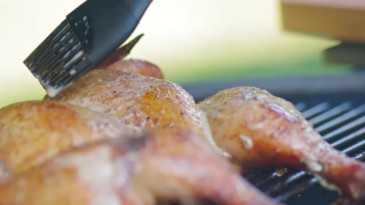 Basting brush spreading white sauce over browned chicken skin on the grill