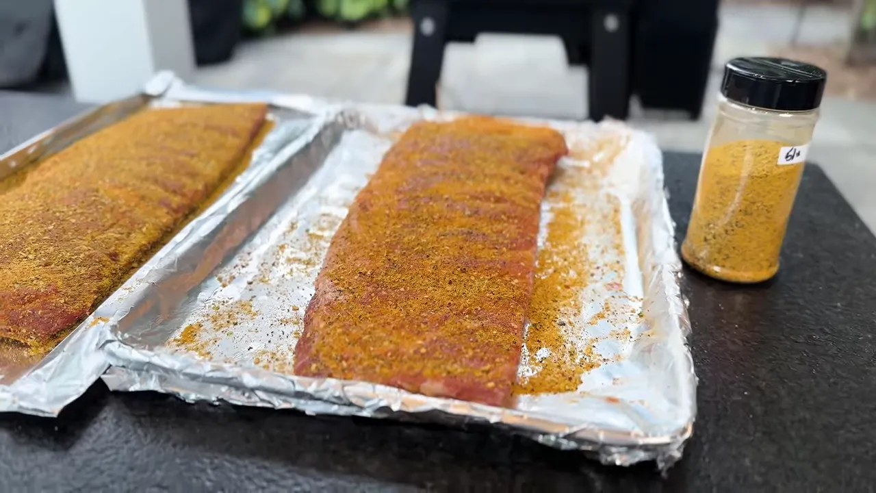 Two seasoned racks of ribs on foil-lined trays beside a yellow rub shaker