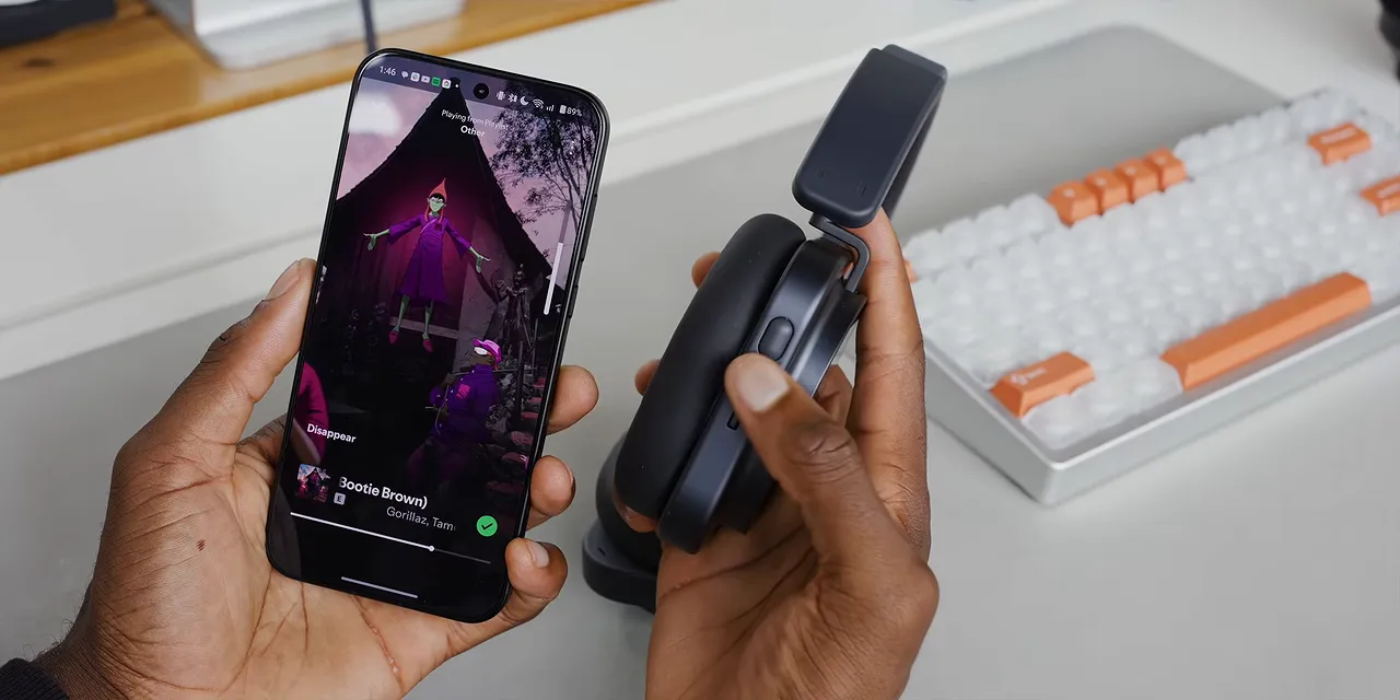 Hand holding a headphone ear cup beside a phone, with the roller and paddle controls visible