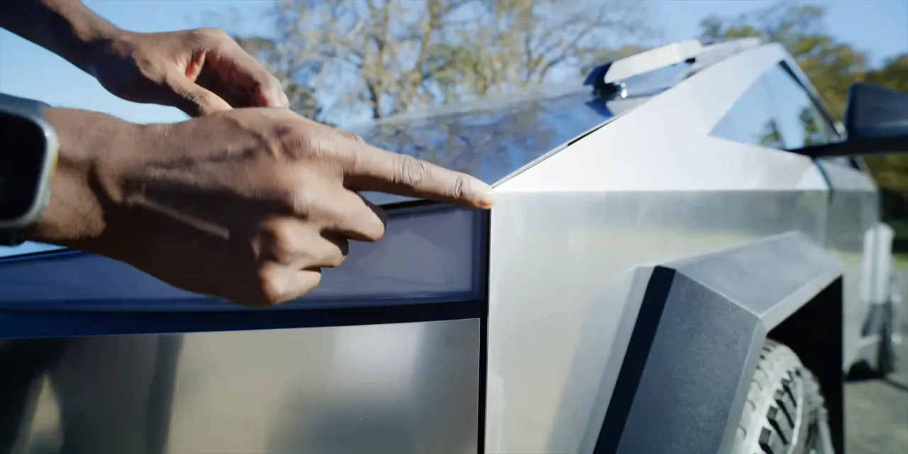 Close-up of a hand pointing at the Cybertruck's angular stainless steel front corner