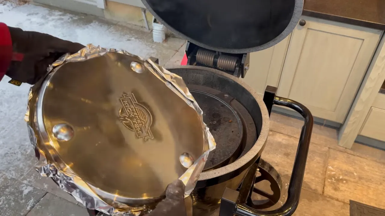 Underside of a round metal drip pan with raised feet held above an open ceramic grill