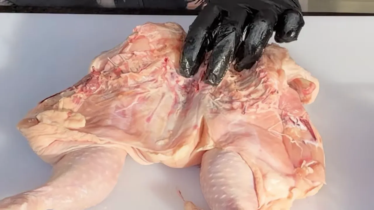 Opened raw chicken with the inner rib cage and central breastbone exposed on a white cutting board