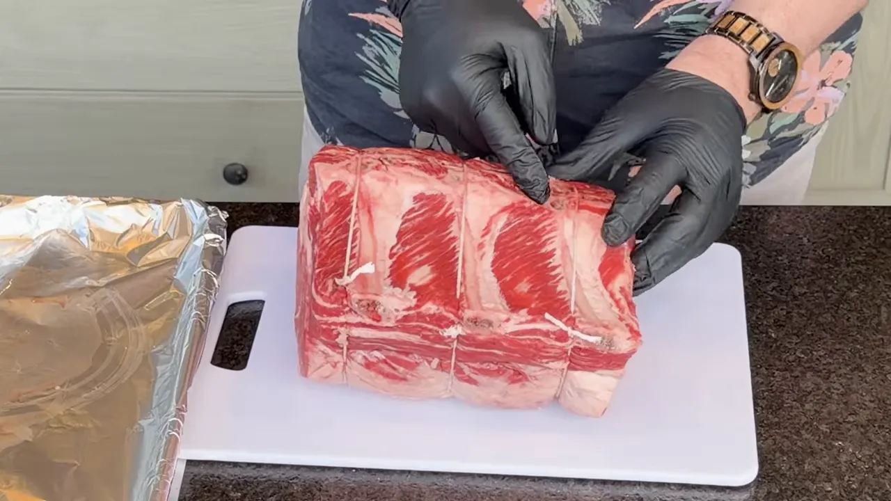 Gloved hands pointing to the bones along the back of a rib roast on a cutting board