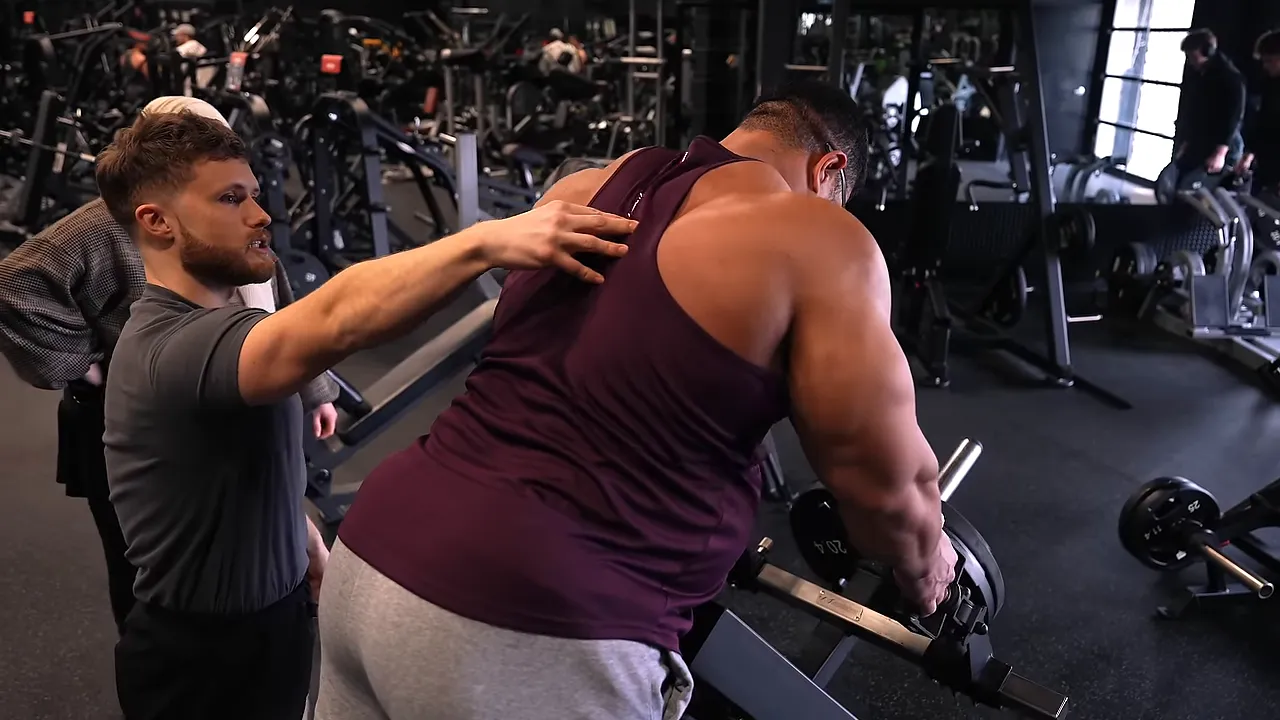 Hassan holds the T-bar row handles with straight arms while Jeff places a hand on his upper back