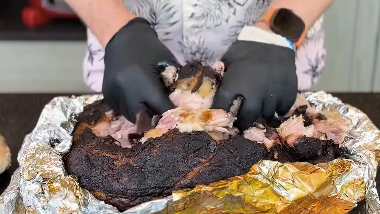 Gloved hands pulling apart pork shoulder to reveal the interior meat beneath dark bark