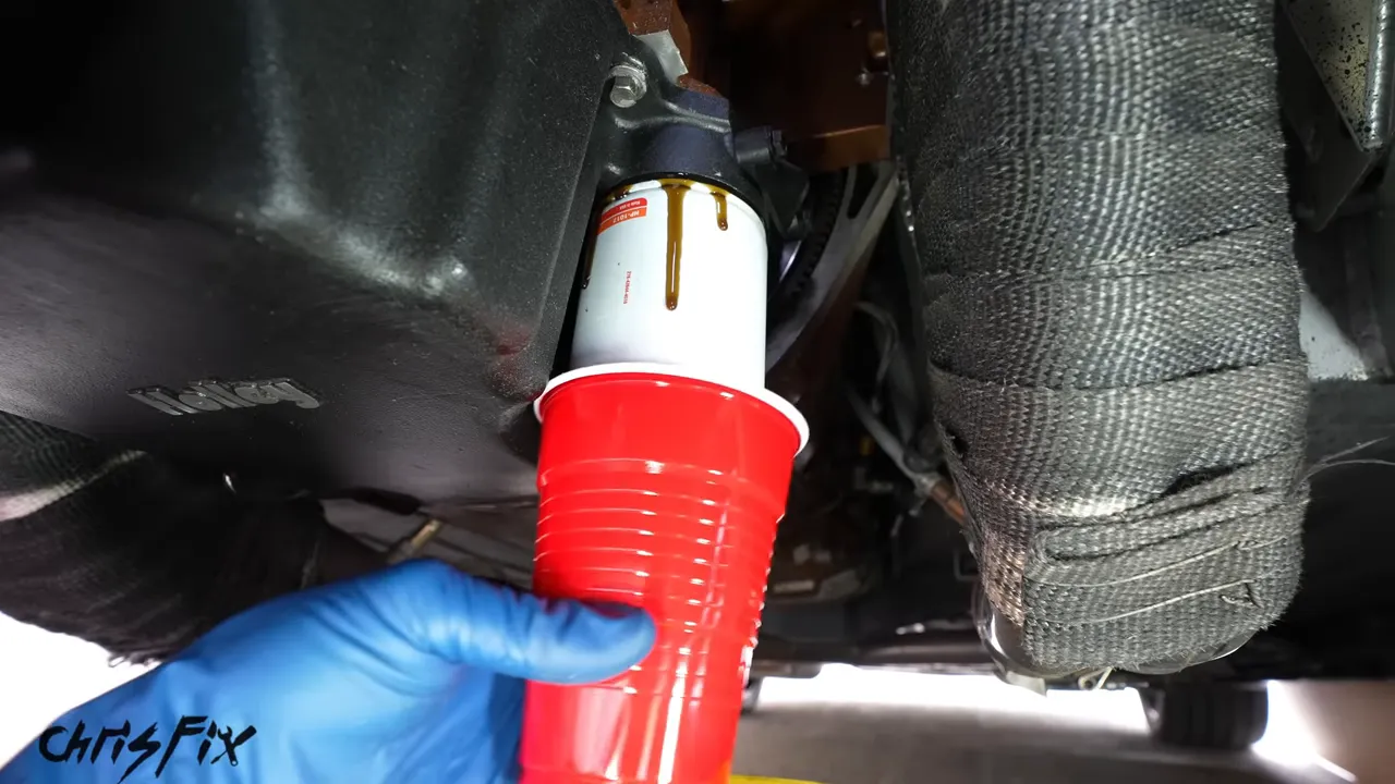 Blue-gloved hand holding a red cup around a vertical oil filter beneath the engine