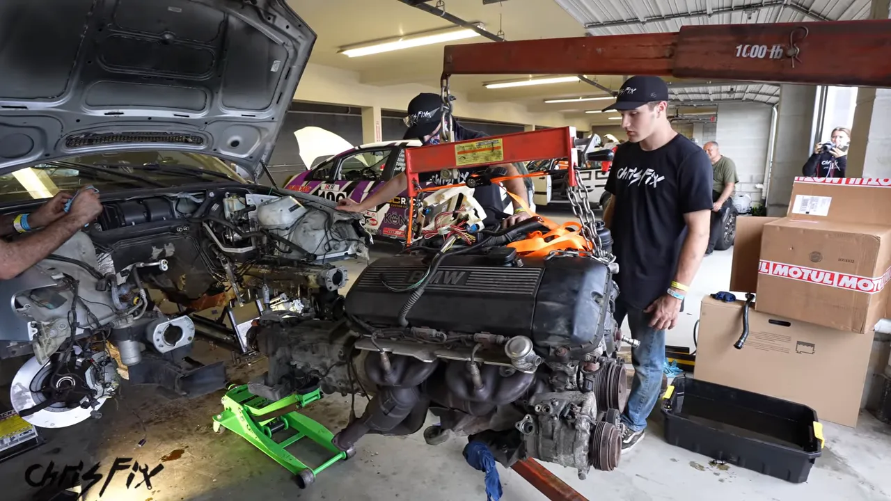 BMW engine and transmission suspended from a hoist in front of the empty engine bay