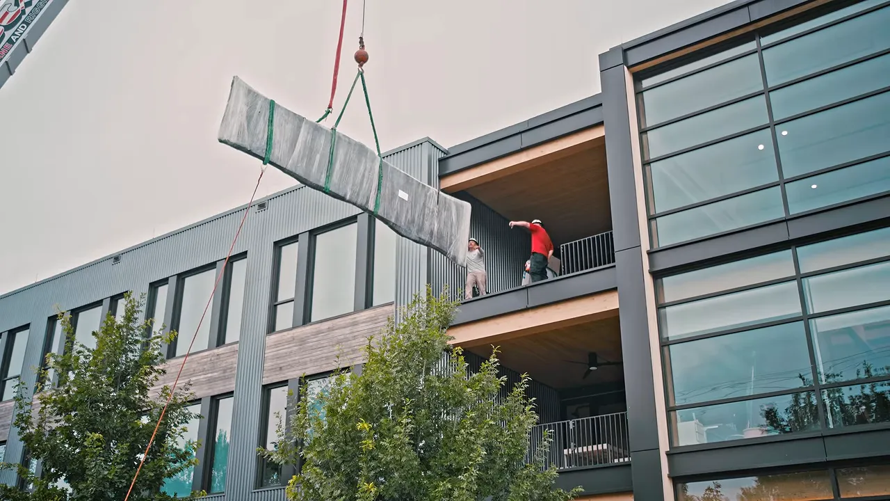 A crane lifting a wrapped tabletop beside an open upper-floor office window