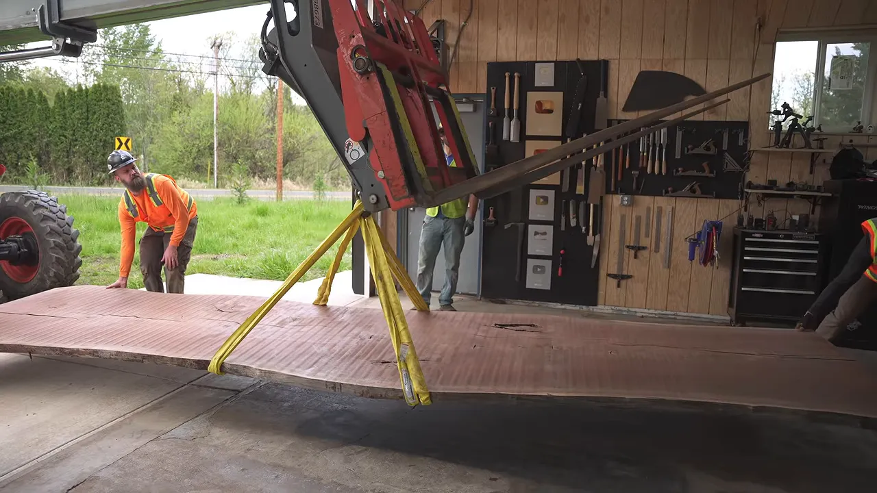 A telehandler and construction crew moving a long walnut slab into the shop