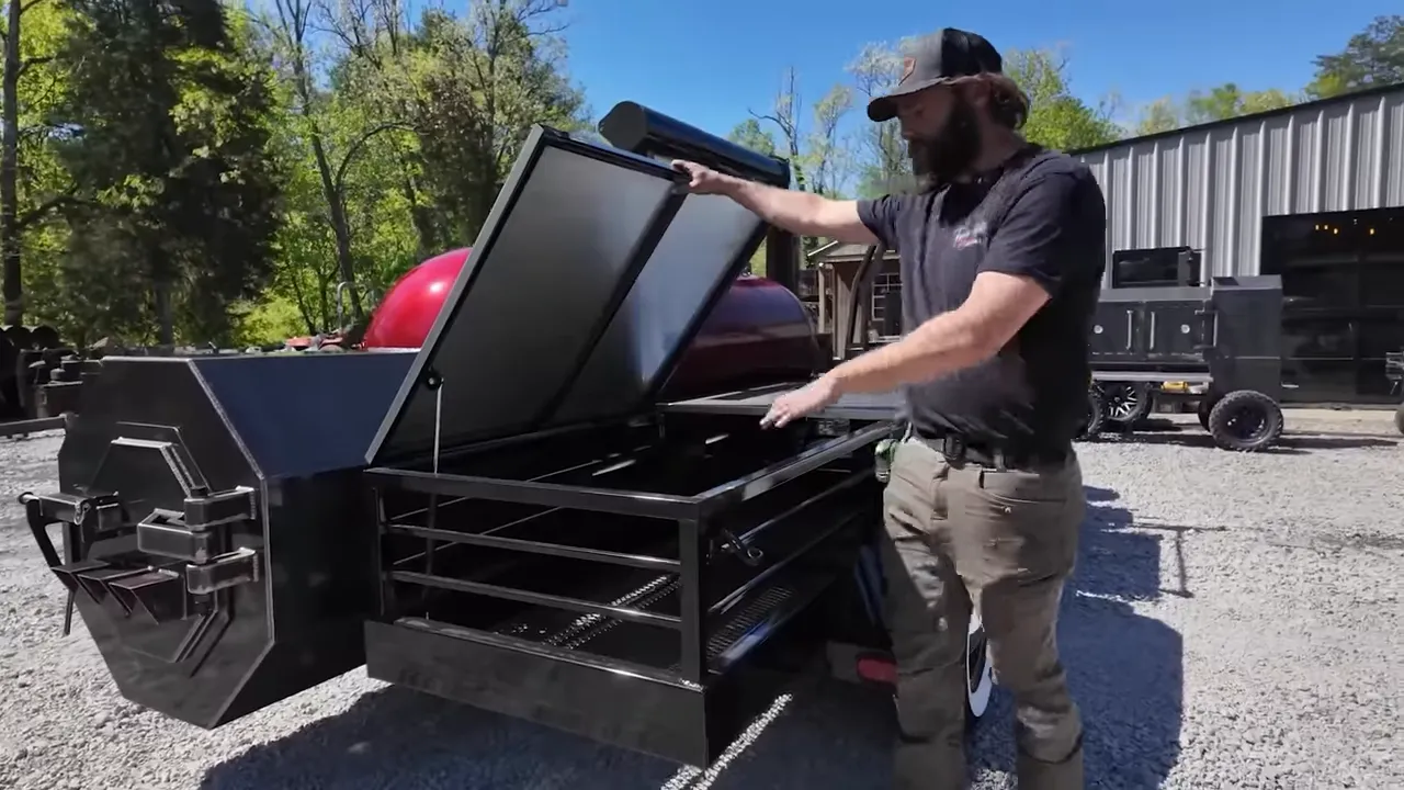 Man holding up a hinged stainless steel shelf beside a red trailer smoker and storage racks