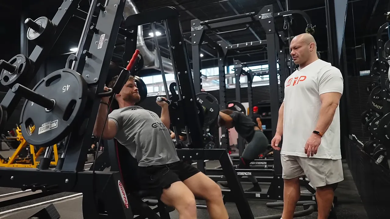 Jeff seated at the bottom of a machine shoulder press with Mike standing beside him