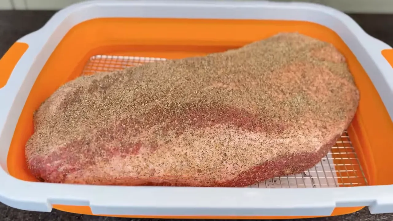 Seasoned brisket resting on a wire rack inside an orange prep tub