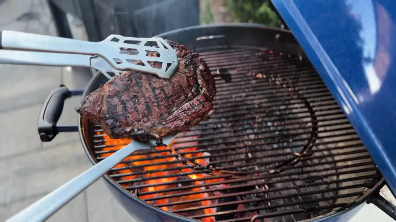 Seared steak held with tongs above glowing coals in a Weber Kettle