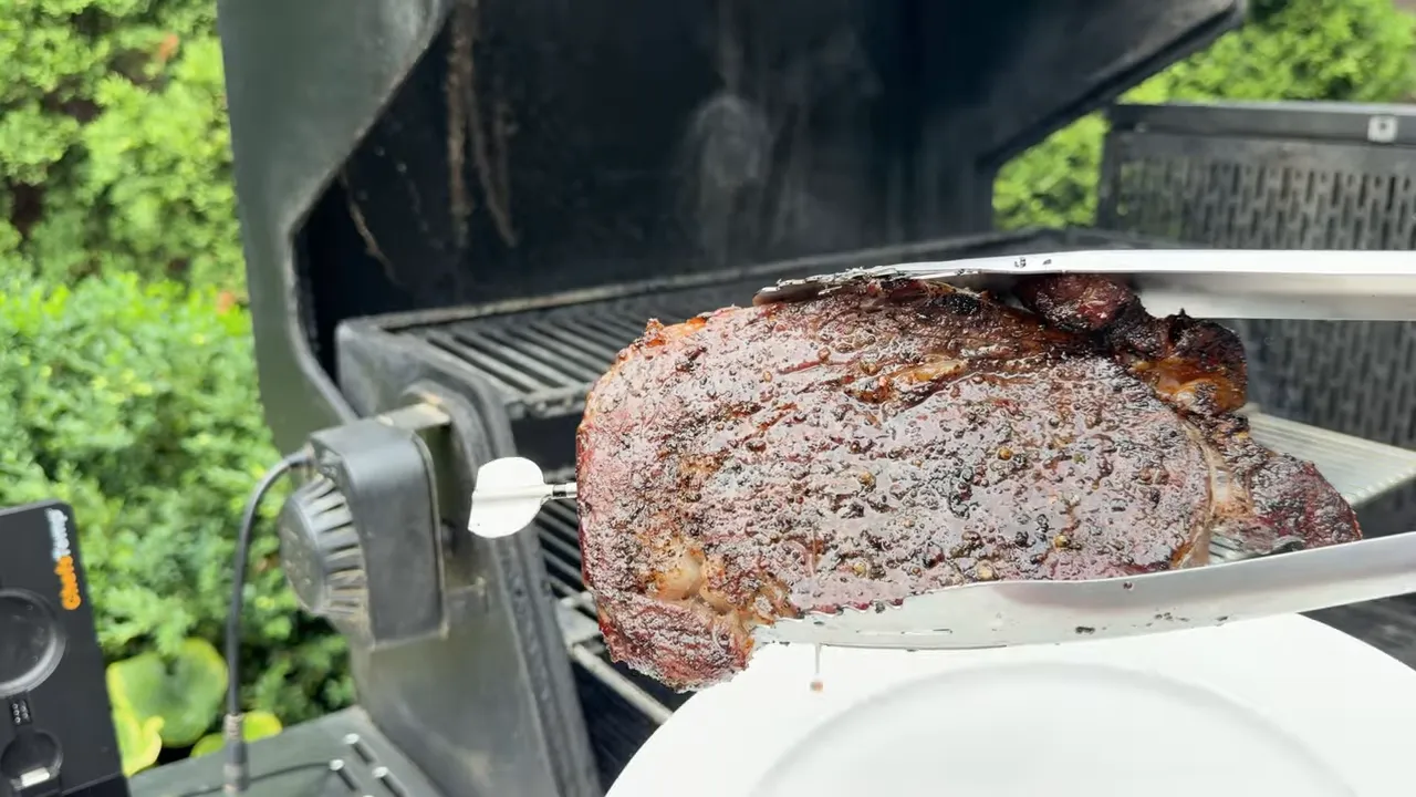 Seared ribeye held in metal tongs above a white plate beside the open grill