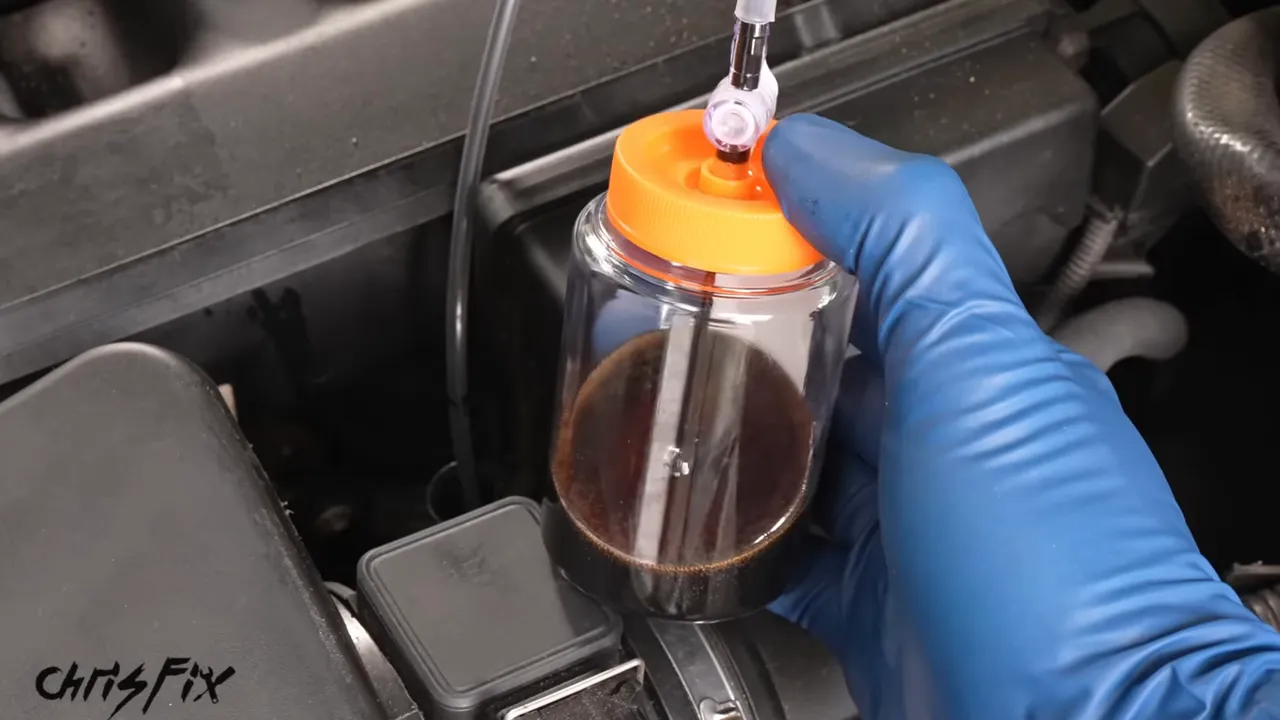 Blue-gloved hand holding a sample bottle filled with dark oil above the engine bay