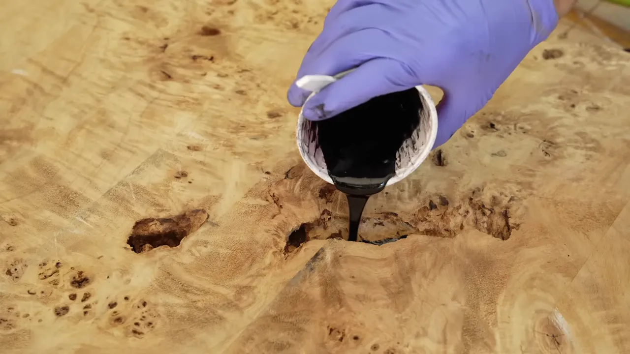 Black epoxy being poured from a cup into a cavity in pale burl wood