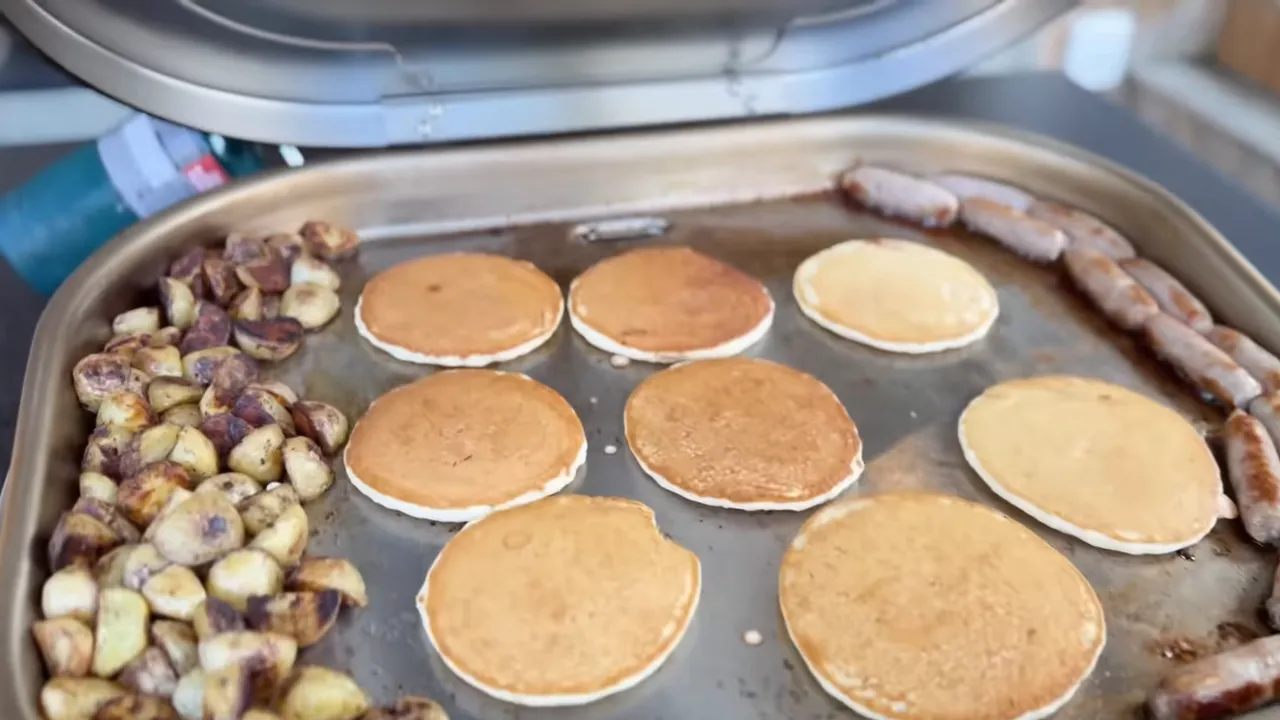 Pancakes cooking in the center of the griddle with potatoes on the left and sausages around the right edge