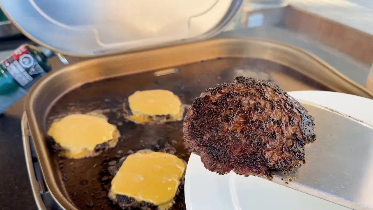 A smash burger held on a spatula showing a dark browned crust, with cheeseburgers on the griddle behind it