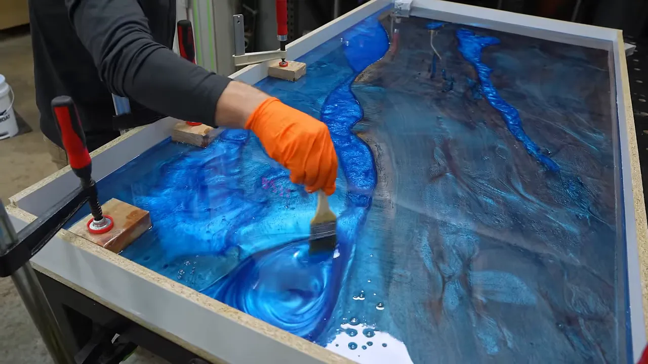 An orange-gloved hand stirring metallic blue epoxy between walnut slabs in a mold