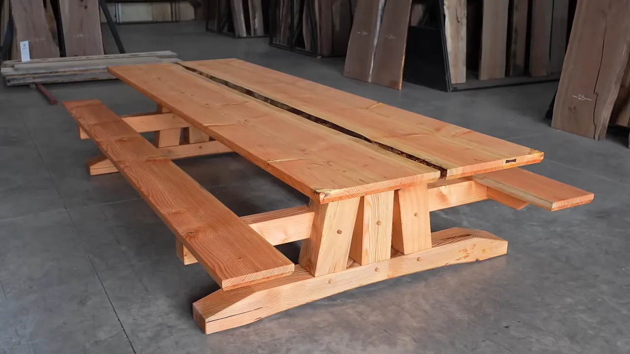 Completed long timber-framed table with attached benches displayed among upright lumber slabs