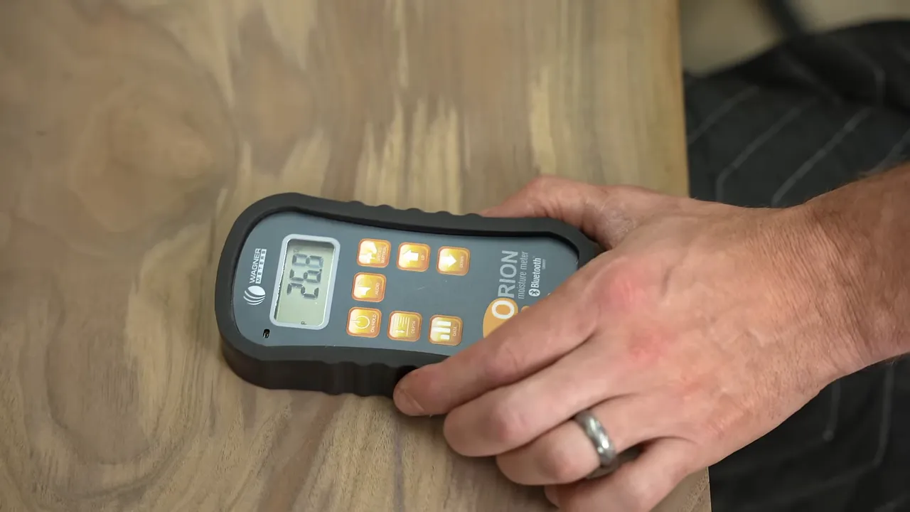 A handheld moisture meter pressed against the surface of a walnut tabletop