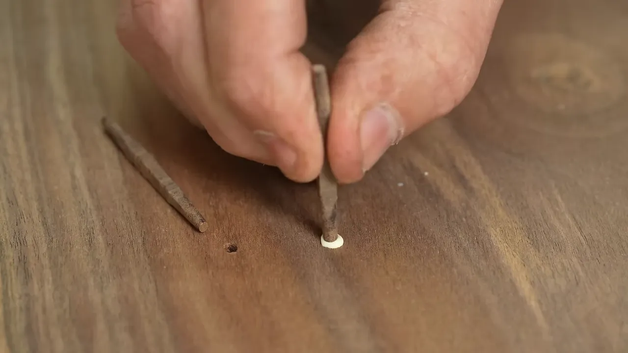Hands positioning slender wooden plugs into small holes in a walnut tabletop