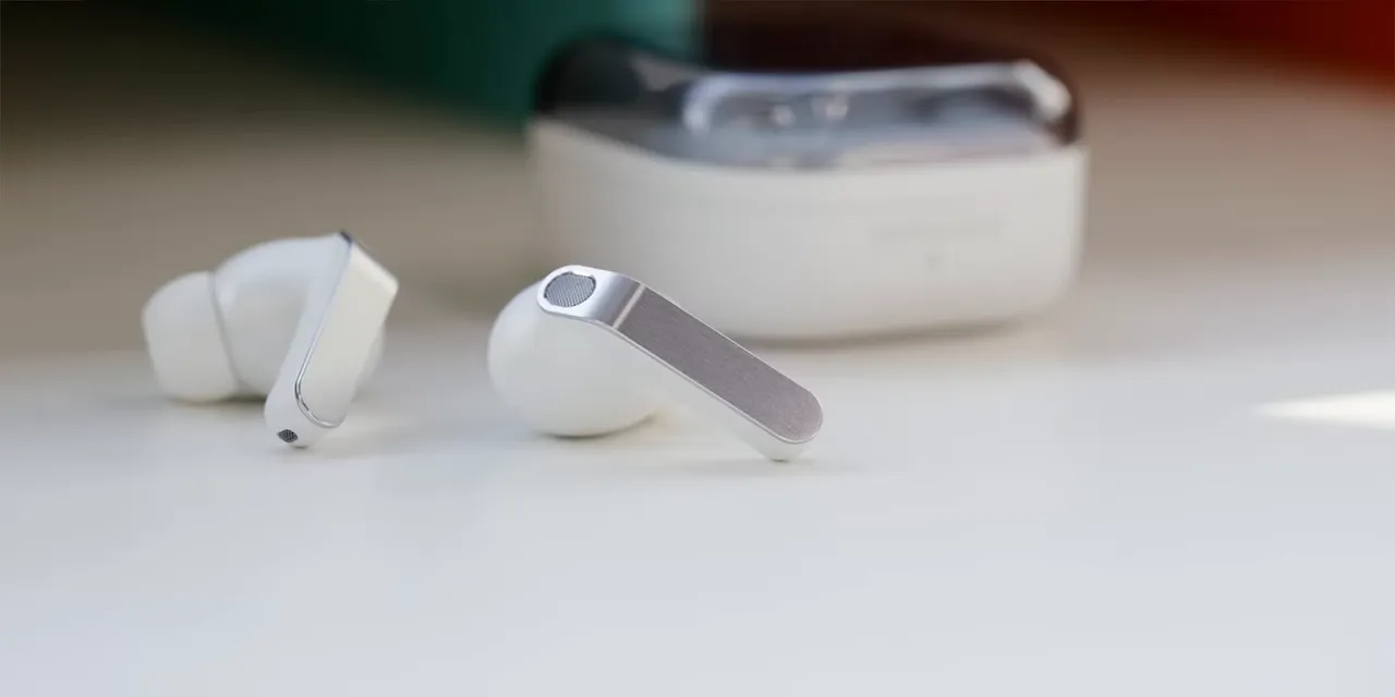 Two white earbuds with flat metallic stems on a desk in front of their closed case