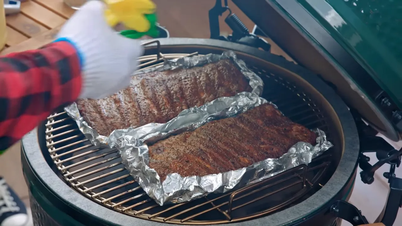 Two rib racks in open-topped foil boats on the Big Green Egg grate