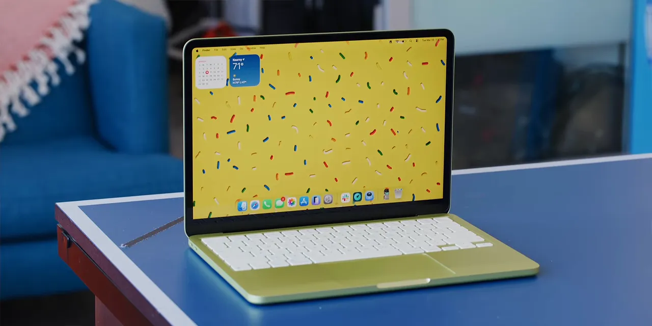 An open yellow-green MacBook Neo on a blue tabletop with a yellow desktop wallpaper