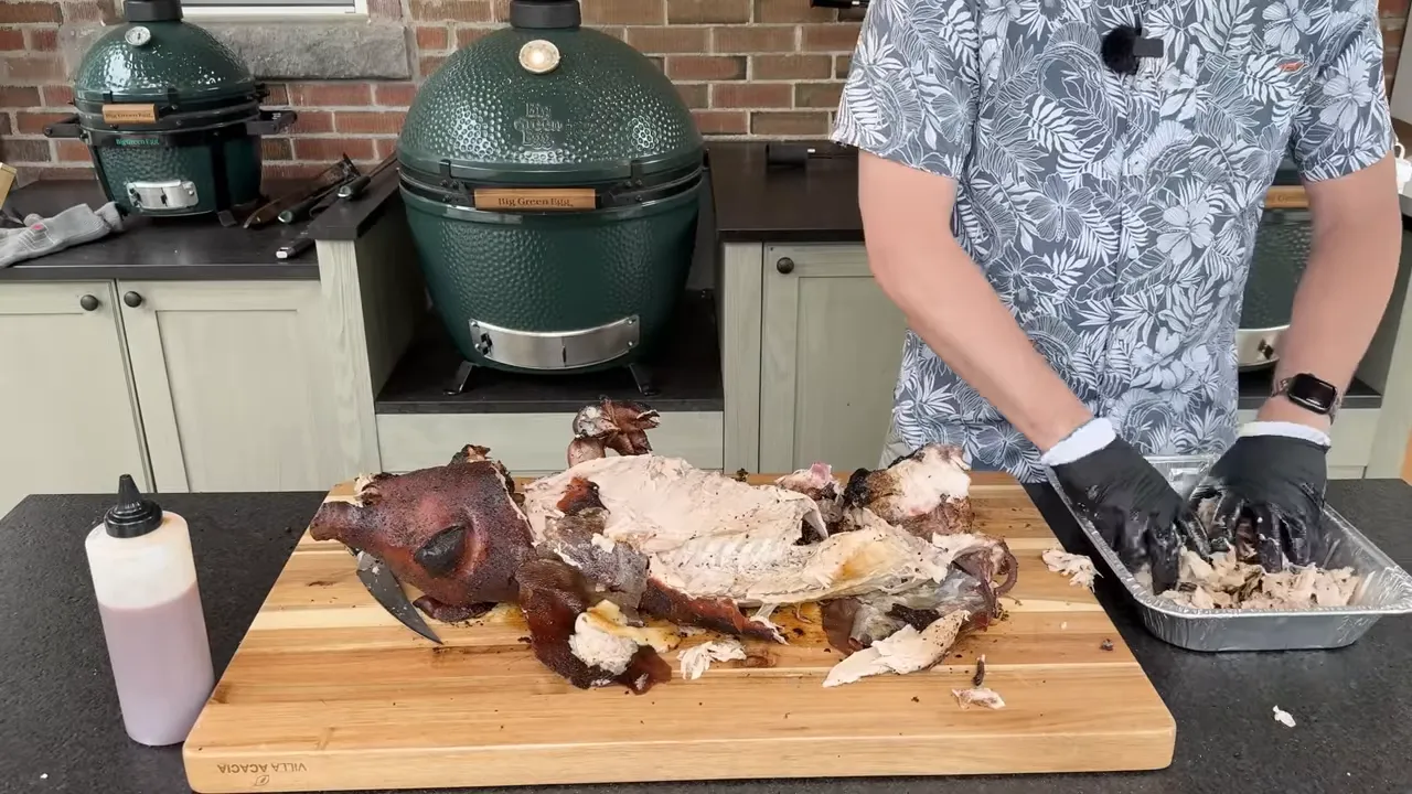 Roasted pig being broken down on a cutting board beside a foil pan of pulled meat