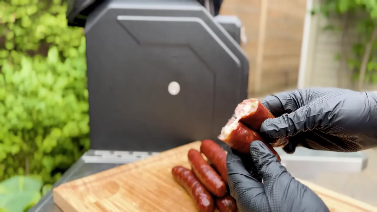 Black-gloved hands holding a broken sausage with its filling exposed above a cutting board
