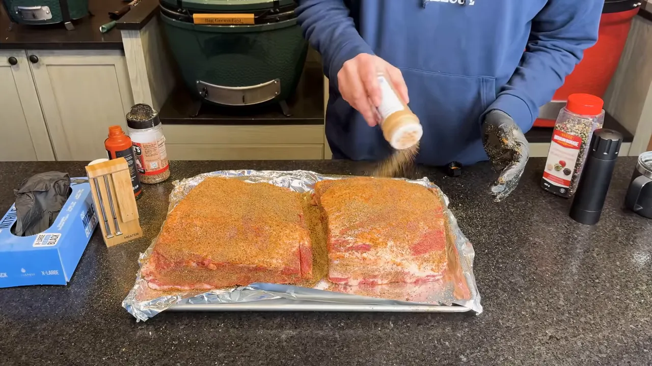 Two seasoned beef rib racks on a foil-lined tray beside seasoning containers