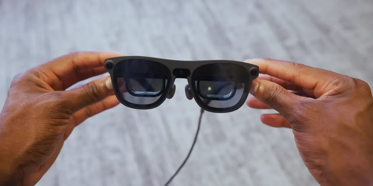 Hands holding Project Aura glasses with cameras visible across the front and a cable hanging below
