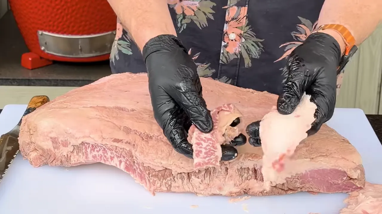 Gloved hands holding pieces of trimmed fat above a raw brisket