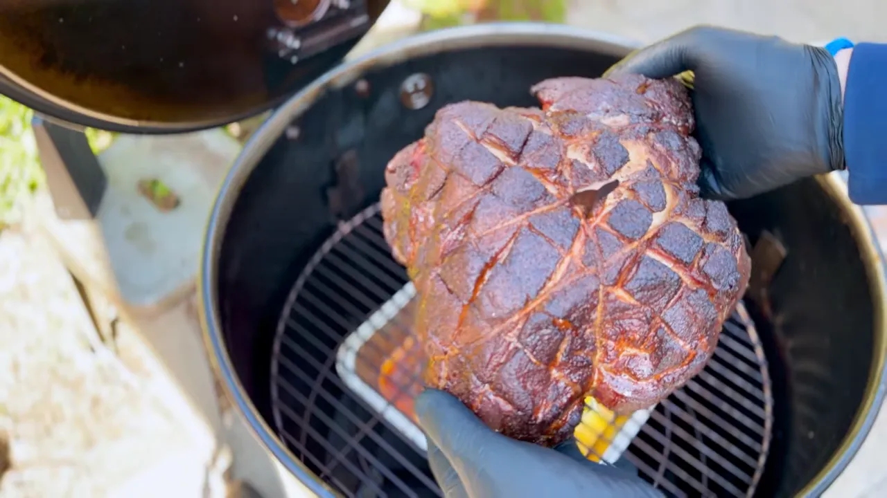 Gloved hand holding a dark-barked pork shoulder above the drum smoker grate