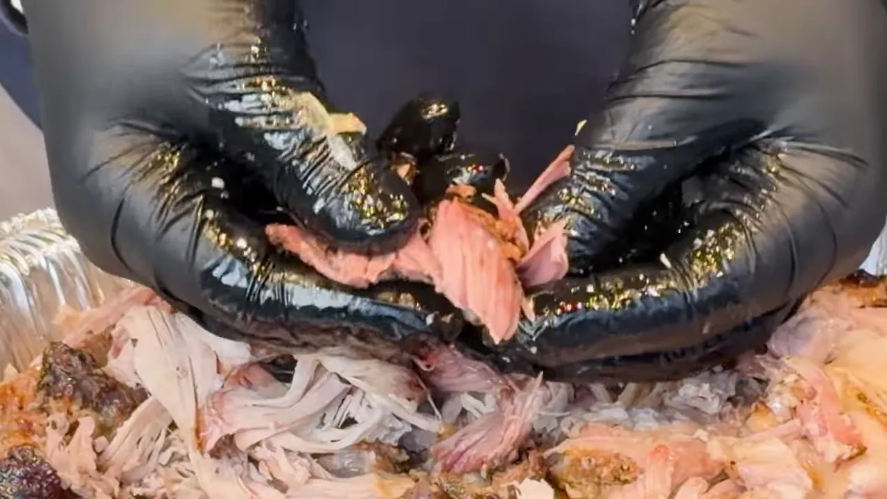 Black-gloved hands pulling apart a piece of pork above a tray of shredded meat