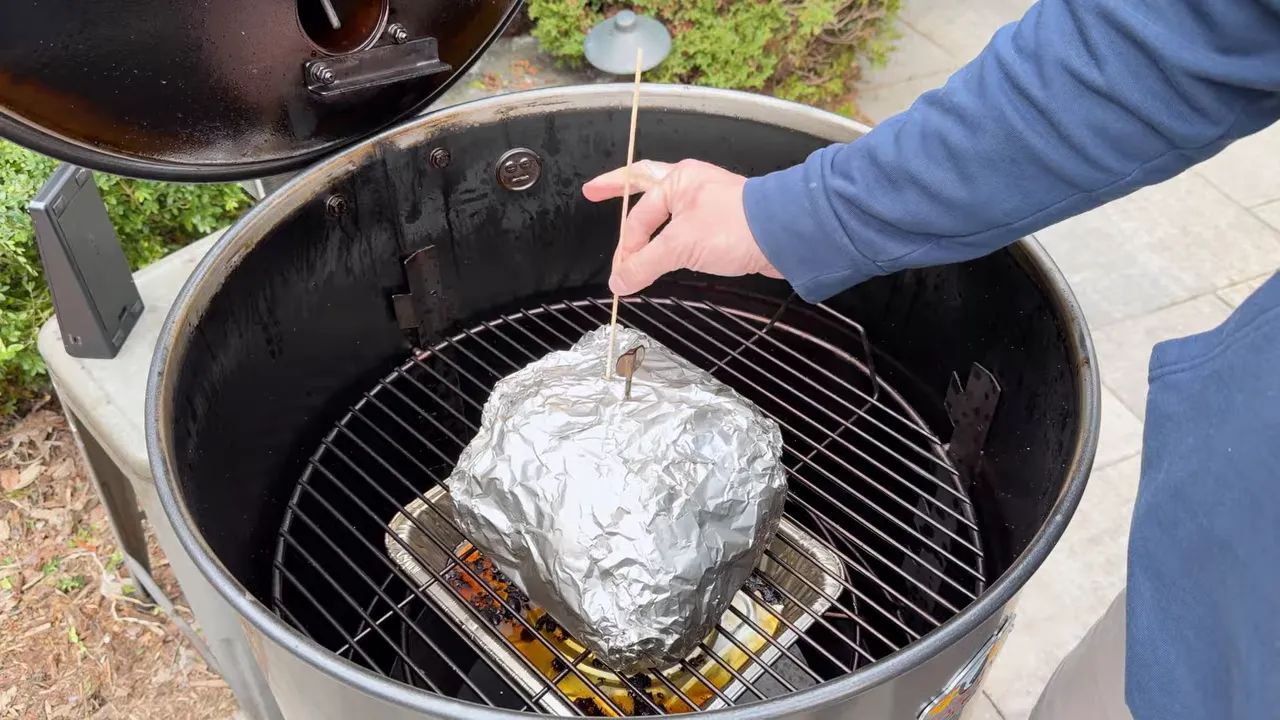 Hand checking a foil-wrapped pork shoulder with a toothpick inside the drum smoker