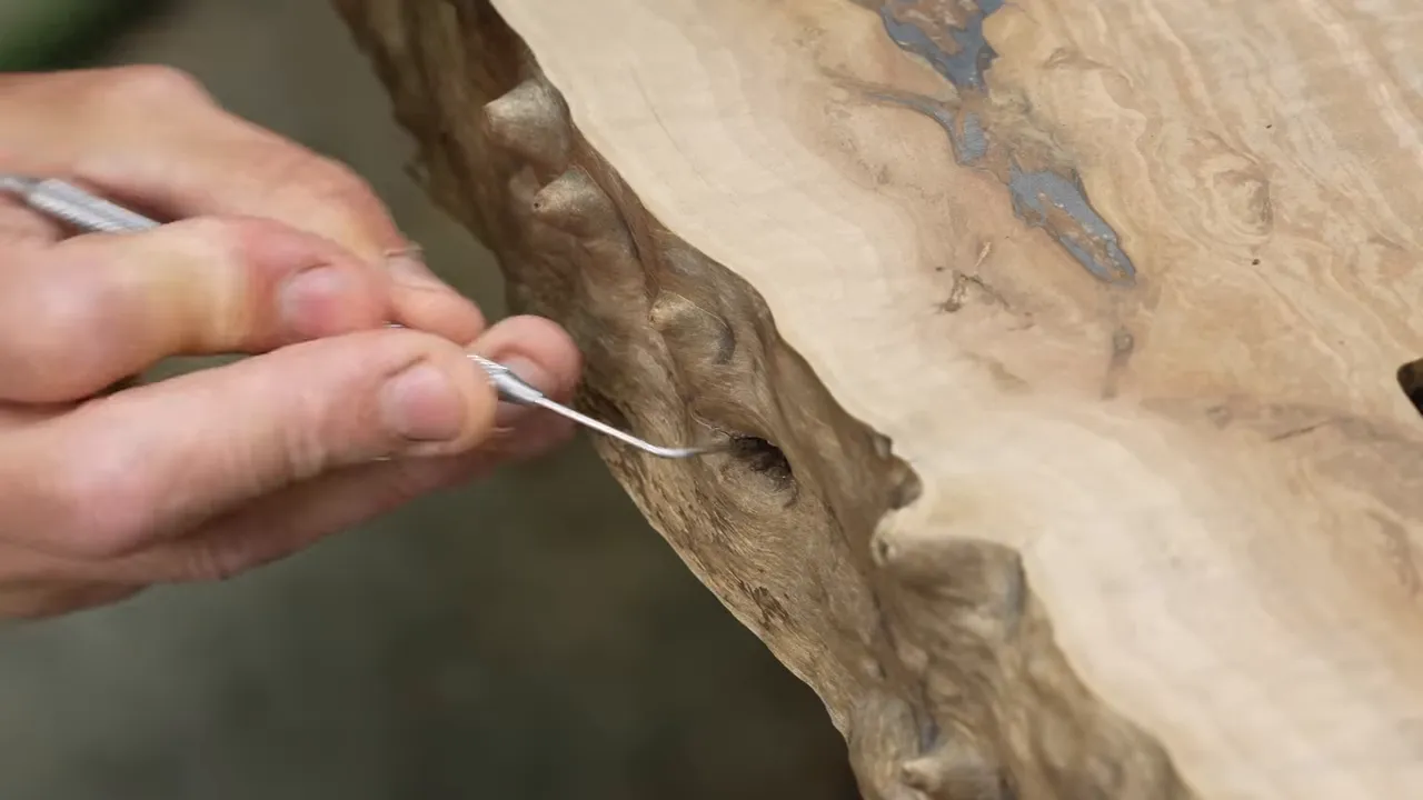 A small metal pick cleaning a recess in the textured live edge of a walnut slab