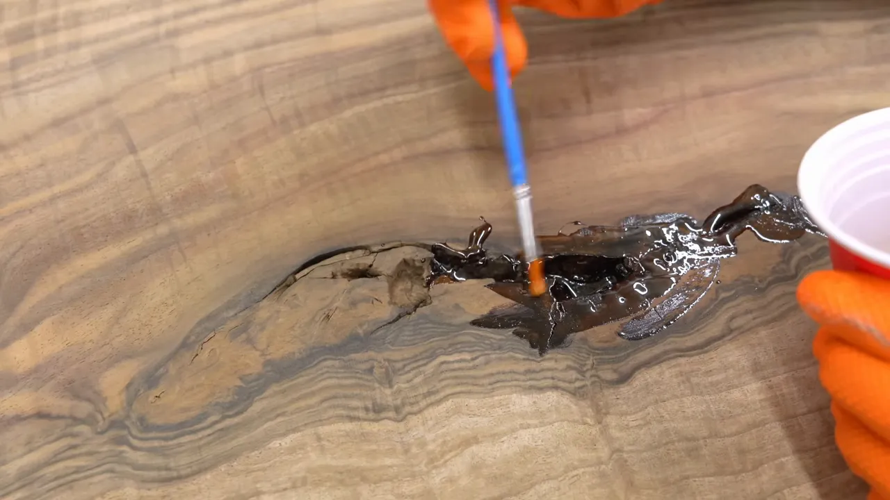 Orange-gloved hands brushing clear resin into a dark void in a walnut slab