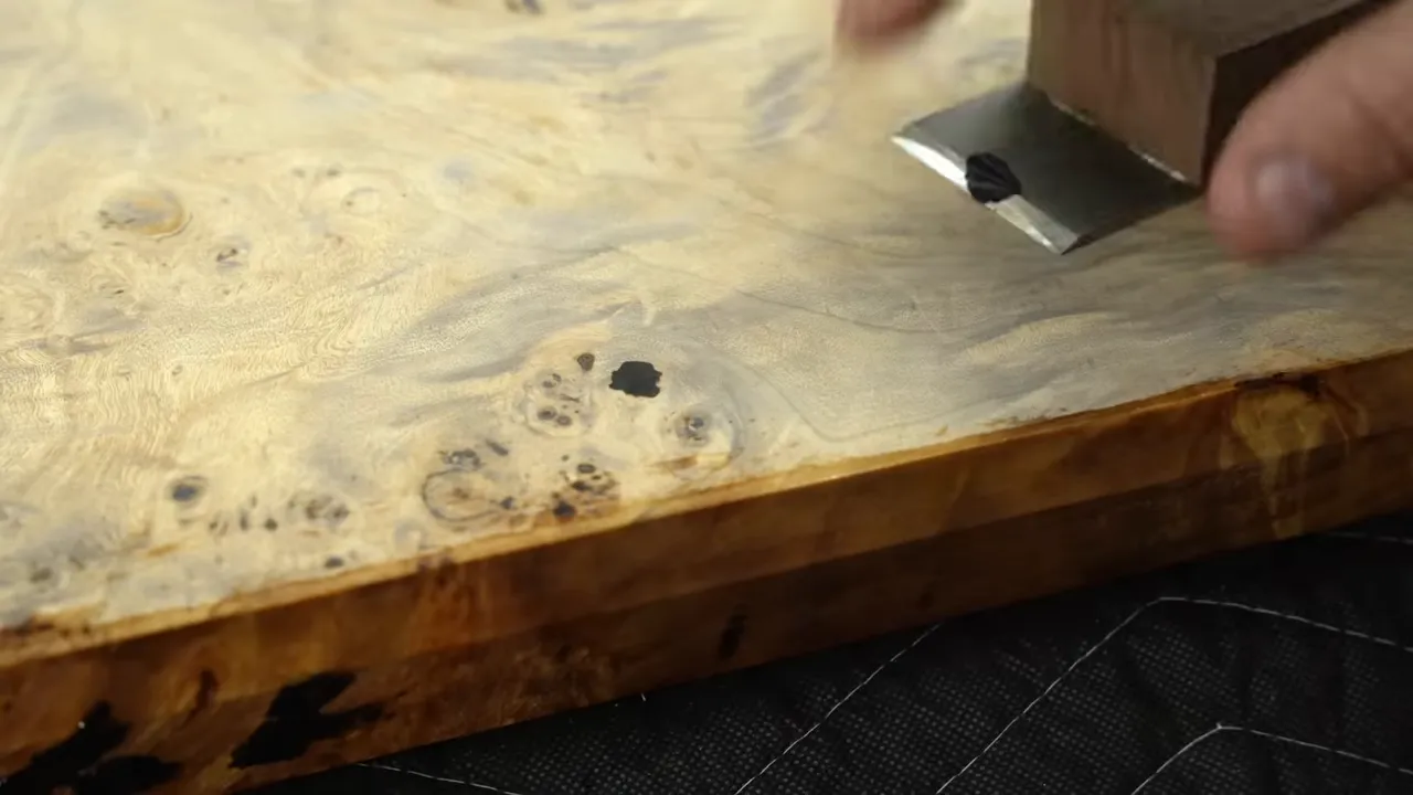 A small scraper leveling dark filler in a pale burl tabletop