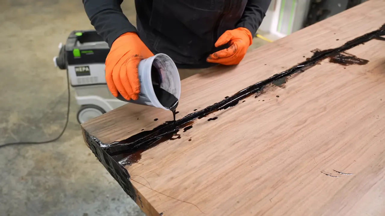 Black epoxy being poured from a mixing cup into a long crack in walnut